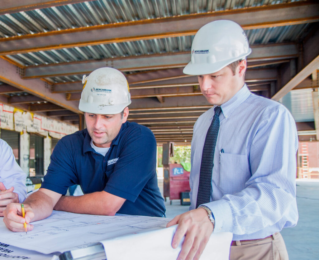 Two Benchmark team members in hard hats review plans and blue prints on a job site in progress